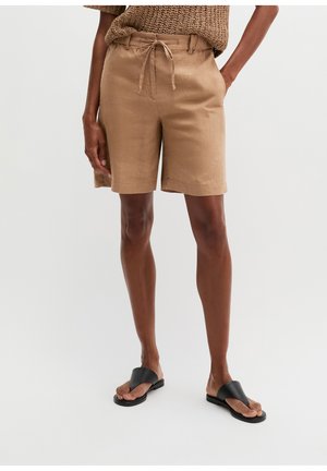 Persoon draagt lichtbruine knielange shorts met trekkoord in de taille, een gebreide top en zwarte sandalen met open teen, staande op een effen achtergrond.