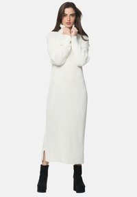 Robe en maille côtelée longue de couleur blanche avec un col haut, dotée de fentes sur les côtés et de manches longues, associée à des bottes à plateforme noires.