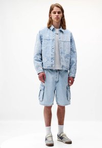 Svetlo modra jeans jakna s teksturiranim vzorcem, v kombinaciji s usklajenimi cargo kratkimi hlačami. Pod njo je bela majica ter teniske z sivimi in modrimi akcenti.