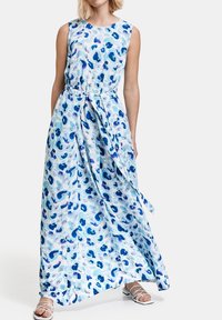 Maxi šaty bez rukávů, z světle modré látky s abstraktními tmavšími modrými a fialovými vzory, stažené v pase zavázaným pásem.
