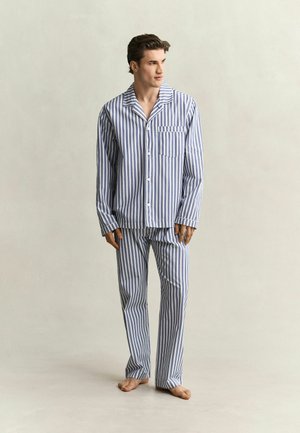 Man die blootsvoets staat en een blauw-wit verticaal gestreept pyjamajasje met knopen en lange mouwen draagt, met bijpassende broek, tegen een effen achtergrond.