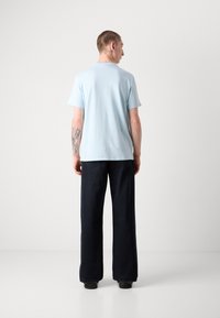 Tricou din bumbac de culoare albastru deschis, cu mâneci scurte, văzut din spate. Asortat cu pantaloni negri largi și un tatuaj discret pe braț.