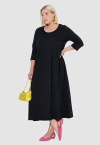 Czarna sukienka z długim rękawem, dopasowaną górą i zwiewnym dołem. Modelka trzyma żółtą torebkę i ma na sobie różowe czółenka.