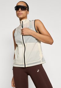 Gilet léger sans manches en maille beige translucide, avec un col montant et une fermeture éclair noire. Associé à des leggings marron portant un logo.