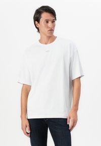 Young man with dark hair wears white oversized t-shirt with small blue logo at chest and dark blue jeans, standing against white background.