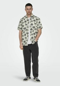 Chemise à manches courtes en crème avec imprimé de palmiers noirs, associée à un pantalon noir et des baskets sombres. Caractérisée par un design décontracté et une coupe relax.