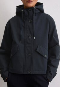 Granatowa kurtka z kapturem wykonana z trwałego materiału, z przodem zapinanym na guziki, dwoma dużymi kieszeniami z przodu oraz regulowanymi sznurkami przy kapturze.