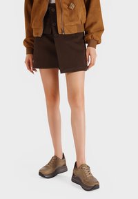Giubbotto bomber in suede marrone con una spilla decorativa, abbinato a una gonna avvolgente marrone scuro e sneakers chunky marroni. Texture liscia in tutto.