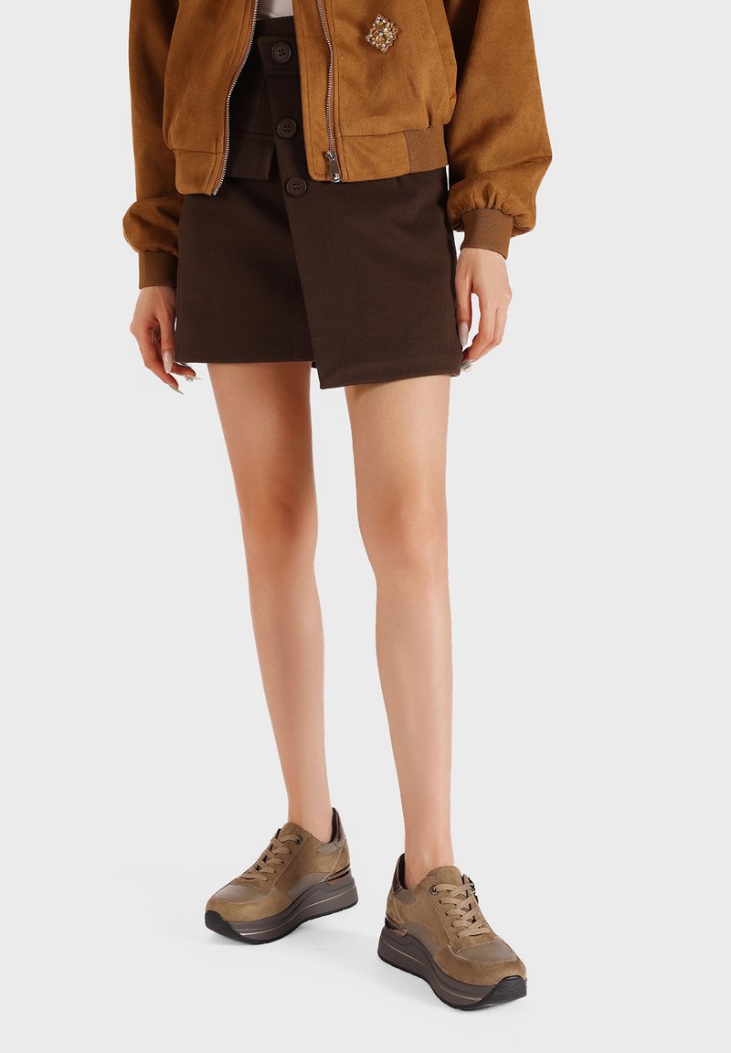 Giubbotto bomber in suede marrone con una spilla decorativa, abbinato a una gonna avvolgente marrone scuro e sneakers chunky marroni. Texture liscia in tutto.