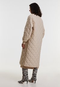 Beige gesteppter langer Mantel mit lockerer Passform und Fledermausärmeln, ausgestattet mit gerippten Manschetten und einer glatten Textur. Getragen mit gestreiften kniehohen Stiefeln.