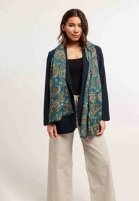 Foulard à motif paisley teal avec bords frangés, superposé sur un haut noir, assorti à un blazer bleu marine et un pantalon large beige.