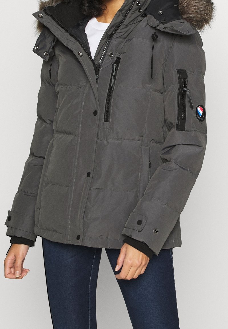 Femme portant une veste d'hiver matelassée grise avec capuche détachable bordée de fourrure, poches zippées sur la poitrine et les manches, et poignets boutonnés.
