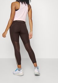 Hnědé legíny s síťovými panely a vysokým pasem, doplněné světle růžovým topem bez rukávů a bílými teniskami s modrými detaily.