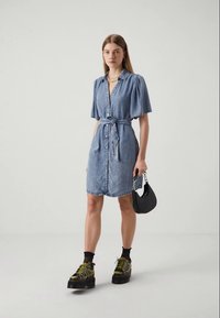 Vestido de camisa em denim azul claro com mangas curtas e volumosas, fecho de botões na frente e cintura atada. Combinado com sapatos de plataforma pretos e chunky e uma bolsa preta.
