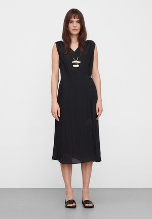 Kobieta w czarnej, bezrękawnej sukience midi, czarnych sandałach typu slide oraz geometrycznym naszyjniku z zawieszką, stojąca na tle jednolitego białego tła.