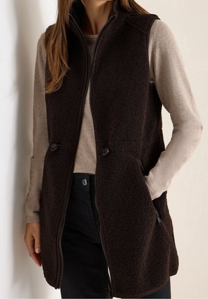 Kobieta ubrana w brązową kamizelkę z polaru sherpa bez rękawów na beżowej bluzce z długim rękawem, z jedną ręką w kieszeni, stojąca przy jasnej ścianie.