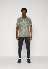 Camisa deportiva en verde y negro con un patrón gráfico, escote redondo y logo. Combinada con pantalones negros ajustados y zapatos negros.