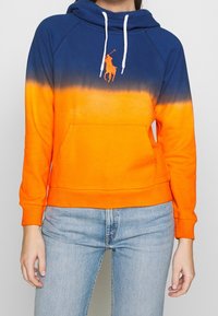 Žena v modro-oranžovém gradientovém mikině s bílým šňůrkami a světle modrých džínách, stojící na jednobarevném pozadí.