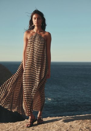 Femme portant une robe rayée fluide à col halter et des sandales, debout sur une falaise rocheuse au bord de la mer sous un ciel dégagé.
