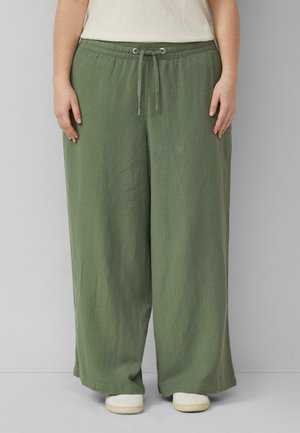 Luźne oliwkowozielone spodnie ze ściągaczem noszone z białymi butami, pokazane od talii do stóp na jasnoszarym tle.