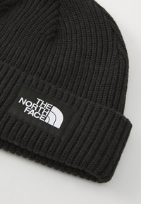 Black knitted beanie with ribbed texture and a folded cuff. Features a rectangular black tag with white text and logo.