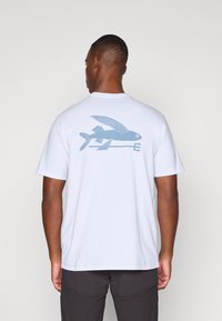 Camisa de algodón blanca con un gráfico azul de un pez volador junto a una flecha, de manga corta y con cuello redondo.