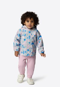 Lätt, mönstrad regnjacka med blå och gröna mönster på en ljus bakgrund, ihop med rosa sweatpants och vita skor.