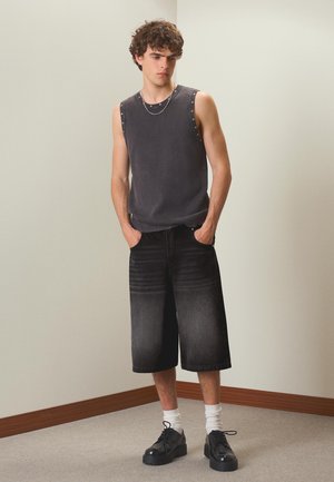 Jonge man met krullend haar draagt een mouwloze donkergrijze top, losse zwarte knielange shorts, witte sokken en zwarte nette schoenen, staat binnen.