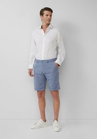 Weißes Leinenhemd, hellblaue gemusterte Shorts mit umgeschlagenen Säumen und weiße Turnschuhe. Das Model steht vor einem schlichten hellgrauen Hintergrund.