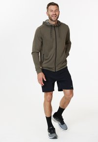 Sweat à capuche zippé vert olive avec cordons, en tissu texturé ; associé à un short noir et des chaussures de sport grises.