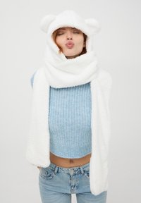 Biała sztuczna futrzana chusta z kapturem i uszami niedźwiedzia, o miękkiej teksturze i szerokim fasonie, w połączeniu z jasnoniebieskim swetrem i jasnymi dżinsami.