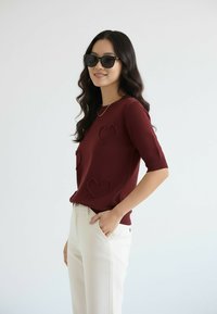 Bordowy dziergany top z akcentami w kształcie serca, zestawiony z białymi spodniami. Okulary przeciwsłoneczne w stylu nowoczesnym i casualowym.