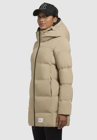 Beige pufferjas met een capuchon, rits aan de voorzijde en zijzakken. Heeft een gestructureerde afwerking en zwarte accenten langs de rits.