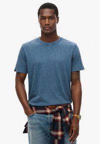 Blå kortærmet t-shirt lavet af blødt stof med rund halsudskæring. En ternet skjorte er bundet rundt om taljen, hvilket fremhæver den afslappede stil.