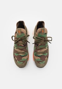 Ett par gröna och bruna kamoflage-sneakers med orange detaljer och snörning i rosetter, sedda ovanifrån på en vit bakgrund.