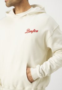 Sudadera beige con un bolsillo frontal y puños acanalados. Presenta el texto "Benfica" bordado en rojo en el lado izquierdo del pecho. Tejido suave y texturizado.