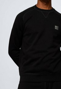 Svart sweatshirt i mjukt tyg, med rundad hals, ribbade ärmslut och en liten präglad logotyp på bröstet.