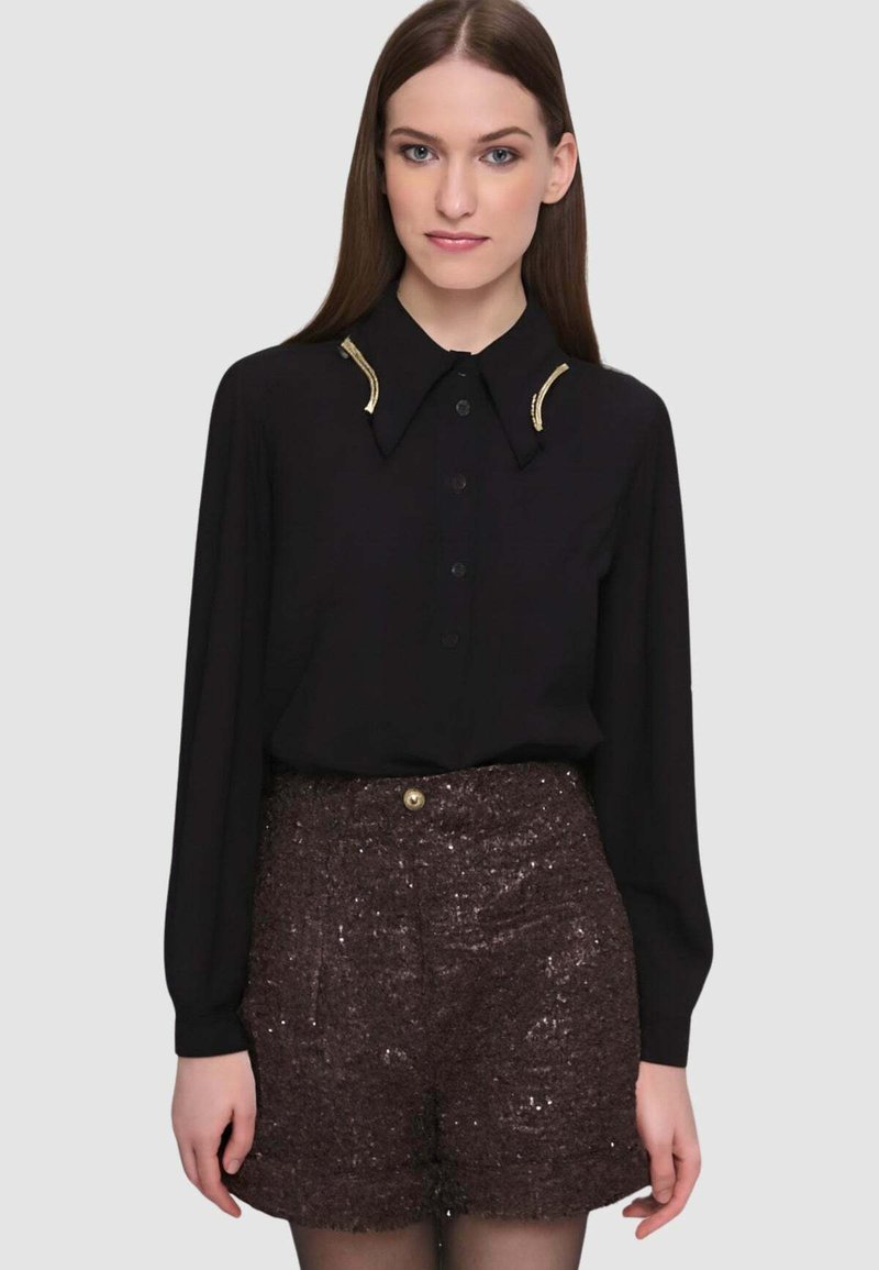 Blusa nera a maniche lunghe con bottoni frontali e dettagli oro sul colletto, abbinata a pantaloncini marroni con paillettes e collant opachi.