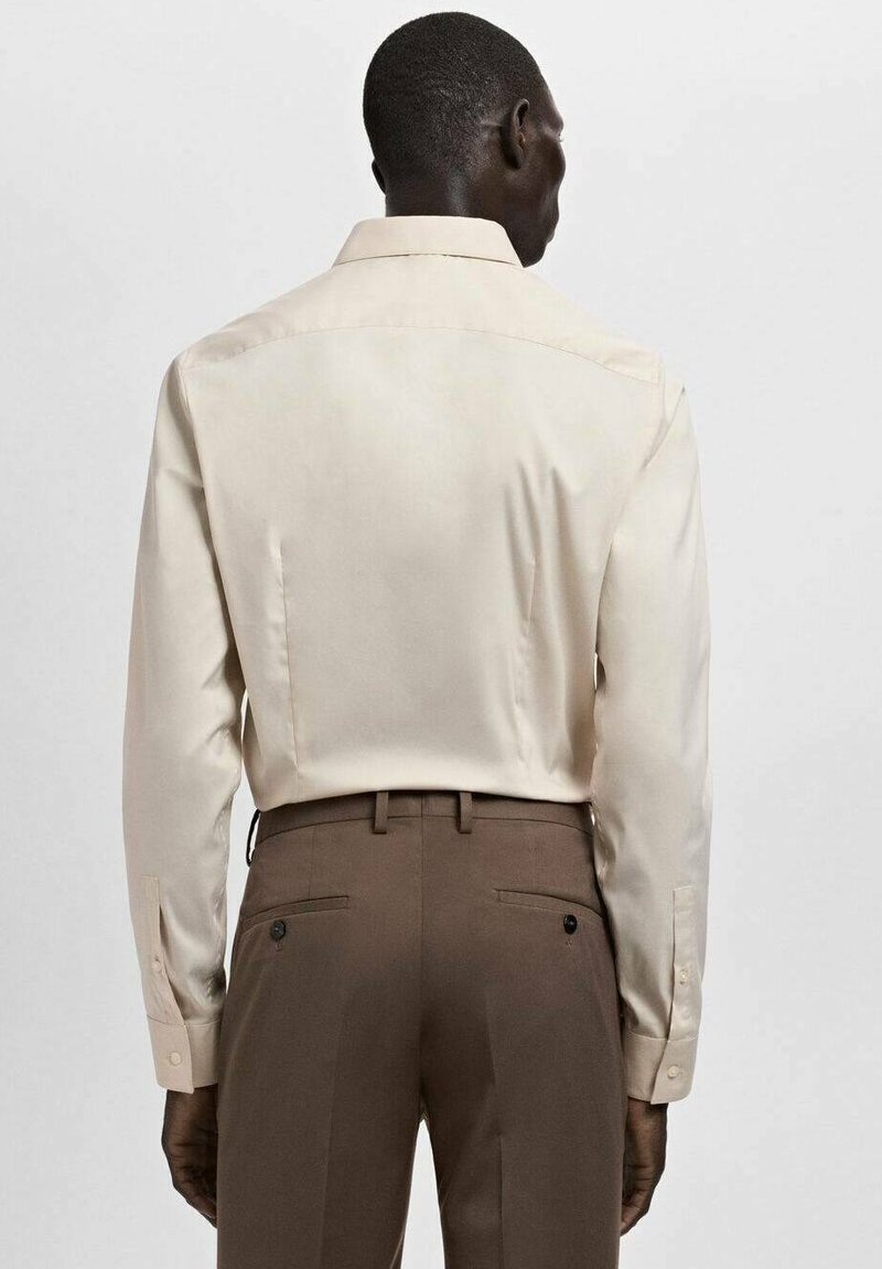 Chemise beige à manches longues avec un col classique, une texture lisse et des poignets à boutons, associée à un pantalon marron avec des poches arrière.