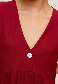 Abito rosso a maniche corte con scollatura a V profonda, vita arricciata e un singolo dettaglio con bottone bianco sul petto. Il tessuto sembra morbido e liscio.