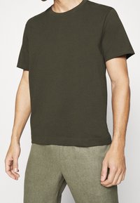 Vihreä oliivinvärinen lyhythihainen t-paita, joka on valmistettu puuvillasta. Paita on pyöreäkauluksinen ja väljä malli, yhdistettynä teksturoituihin beigeihin housuihin.