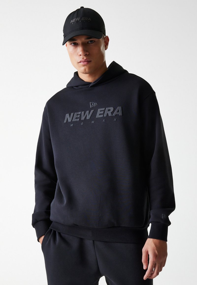 Felpa nera con cappuccio e tasca a marsupio frontale, con un logo "NEW ERA" in rilievo. Indossata con un berretto nero abbinato e pantaloni.