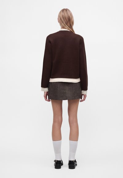 Femme aux longs cheveux blonds portant un pull marron foncé, une jupe à fines rayures, des chaussettes blanches et des chaussures noires, debout de dos sur fond blanc.