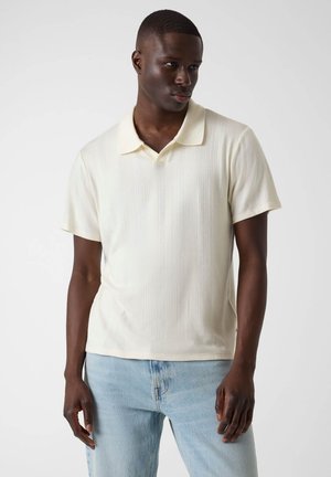 Homme portant un polo crème à manches courtes et un jean bleu clair, debout devant un fond uni clair.