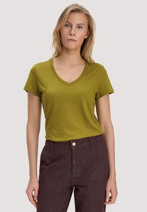 Blonde vrouw staand, gekleed in een olijfgroen T-shirt met V-hals en korte mouwen en donkerbruine, hoogwaterbroek tegen een effen achtergrond.