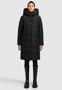 Zwarte donsjas met capuchon, ritssluiting aan de voorkant en doorgestikt patroon. De jas is kniehoog met zijzakken, gecombineerd met kniehoge zwarte laarzen.