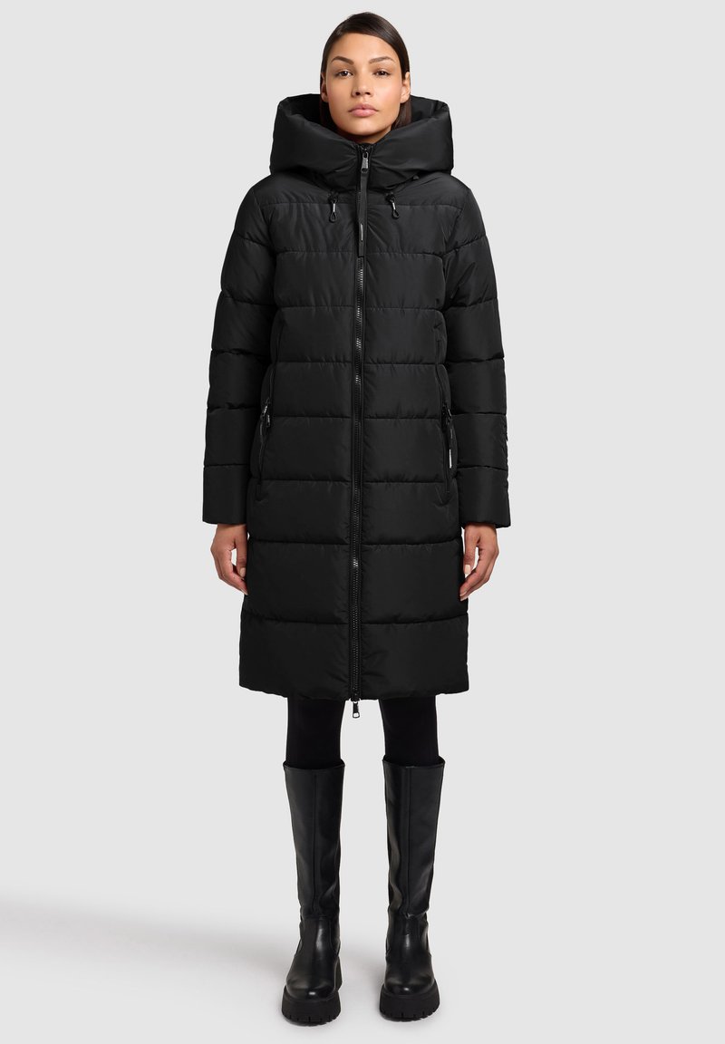 Zwarte donsjas met capuchon, ritssluiting aan de voorkant en doorgestikt patroon. De jas is kniehoog met zijzakken, gecombineerd met kniehoge zwarte laarzen.