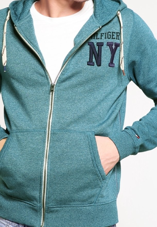 Homme portant un sweat à capuche zippé bleu sarcelle avec les lettres "NY" sur la poitrine, des cordons blancs, et les mains dans les poches avant, par-dessus un t-shirt blanc.