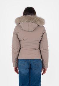 Beżowa kurtka puchowa z kapturem wyłożonym futrem. Ma dopasowany krój, długie rękawy i gładką fakturę. Noszona z niebieskimi dżinsami.