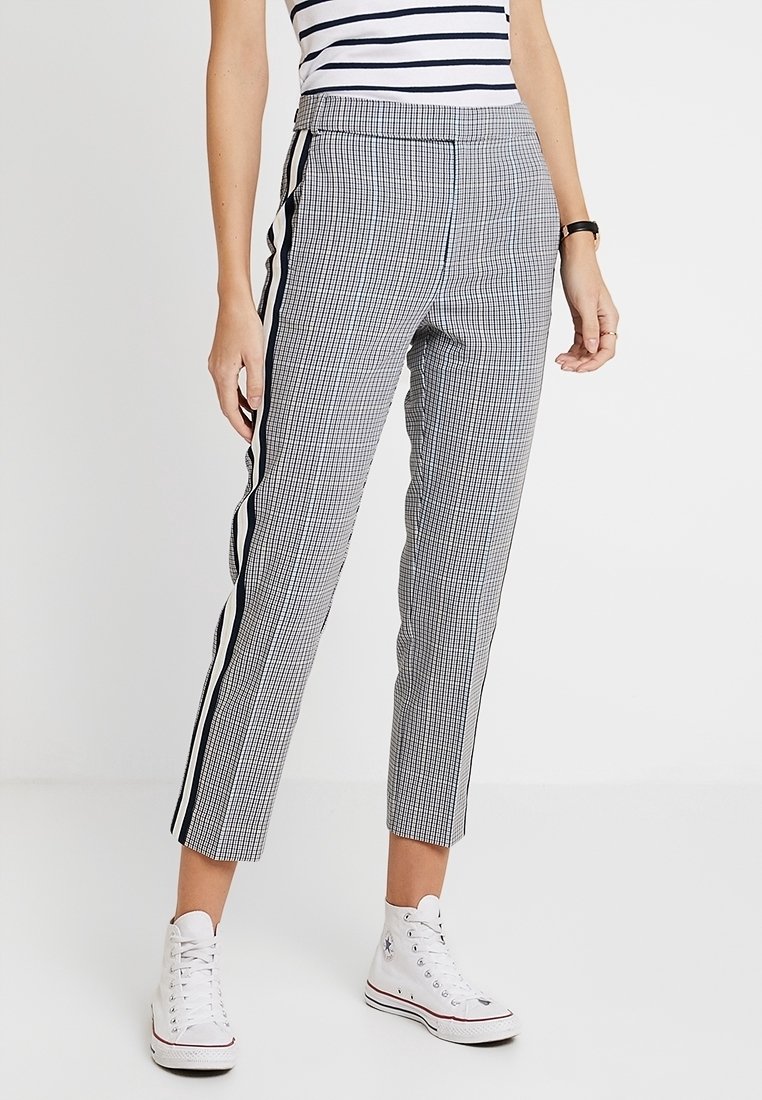 Pantalons à carreaux noir et blanc avec jambe fuselée, rayures sur les côtés et taille haute, fabriqués dans un tissu lisse, associés à des baskets blanches.